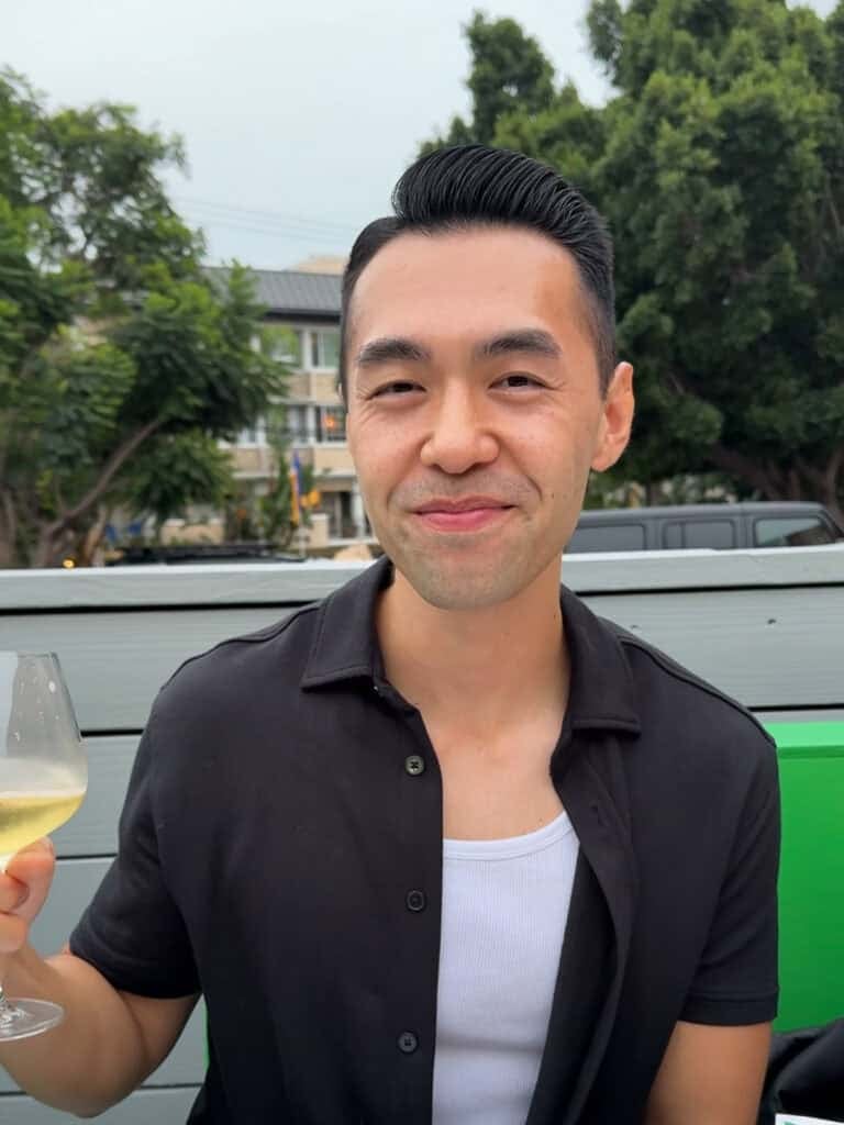 Herman smiling in a black button-down shirt and white undershirt holds a glass of white wine outdoors, featured in his about me portrait.