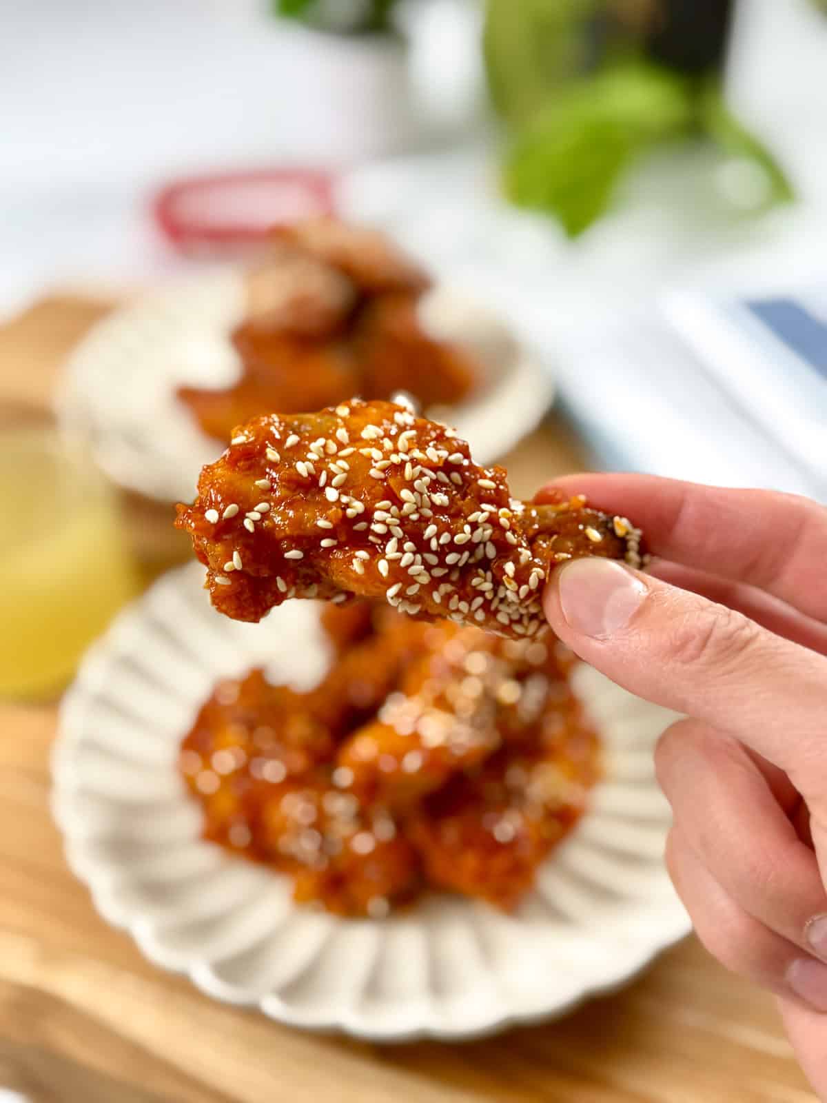 A hand holds a piece of fried chicken coated in a sticky sauce and sprinkled with sesame seeds. In the background, more chicken pieces on a white plate and a drink are visible on a wooden surface.