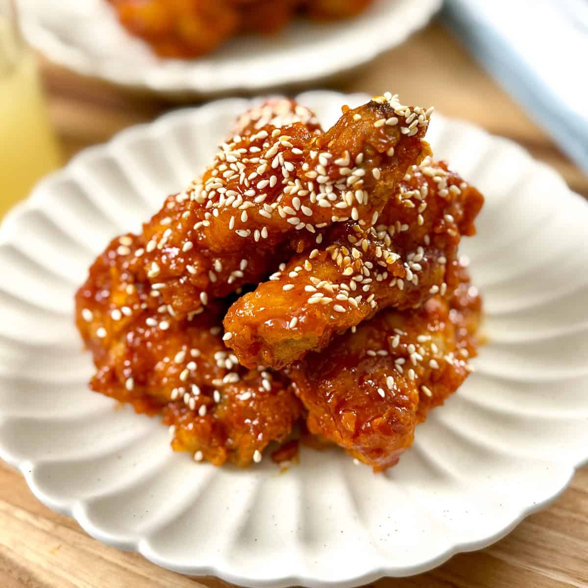 A plate of crispy Korean fried chicken wings coated in a sticky, bright red sauce, garnished with white sesame seeds, served on a scalloped white dish.