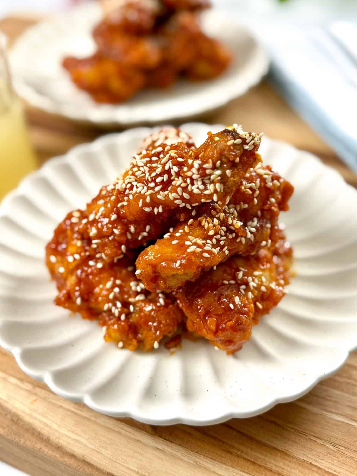 A plate of crispy fried chicken wings coated in a sticky red sauce and sprinkled with sesame seeds, served on a white scalloped dish with more wings in the background.
