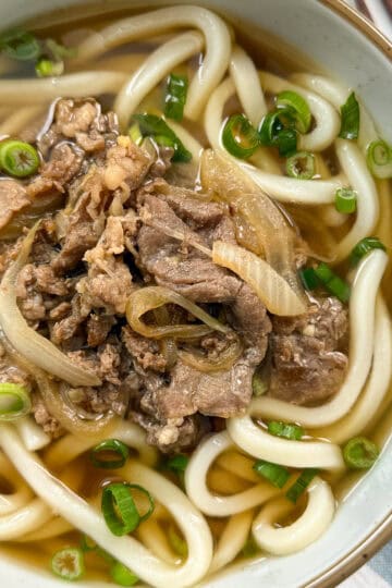 A bowl of udon noodle soup topped with sliced beef, onions, and chopped green onions, with a clear broth. Chopsticks are partially visible at the side.