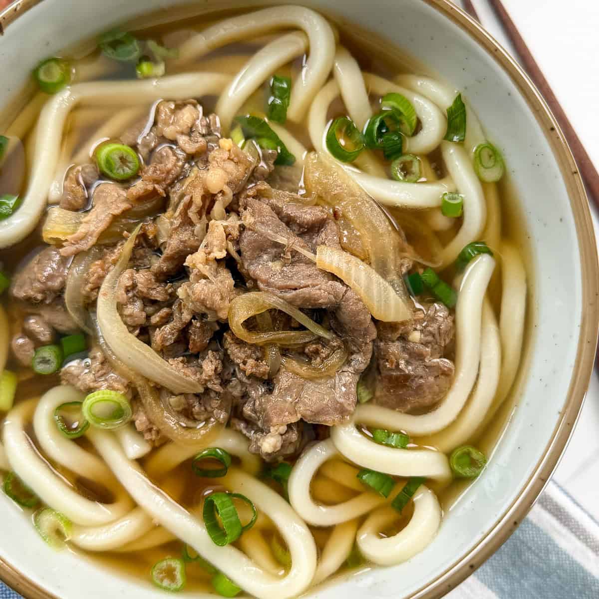 A bowl of udon noodle soup topped with sliced beef, onions, and chopped green onions, with a clear broth. Chopsticks are partially visible at the side.