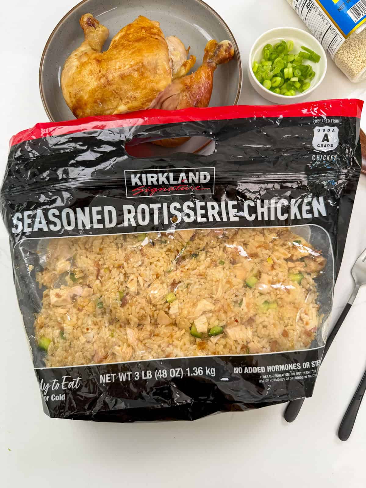 A bag of Kirkland Signature seasoned rotisserie chicken sits on a white surface, with shredded chicken inside. In the background are a plate with cooked chicken, a bowl of sliced green onions, and utensils.