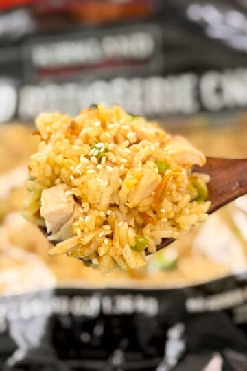 A wooden spoon holds a close-up of costco chicken scallion rice with chicken, vegetables, and sesame seeds, with a blurred bag of frozen fried rice in the background.