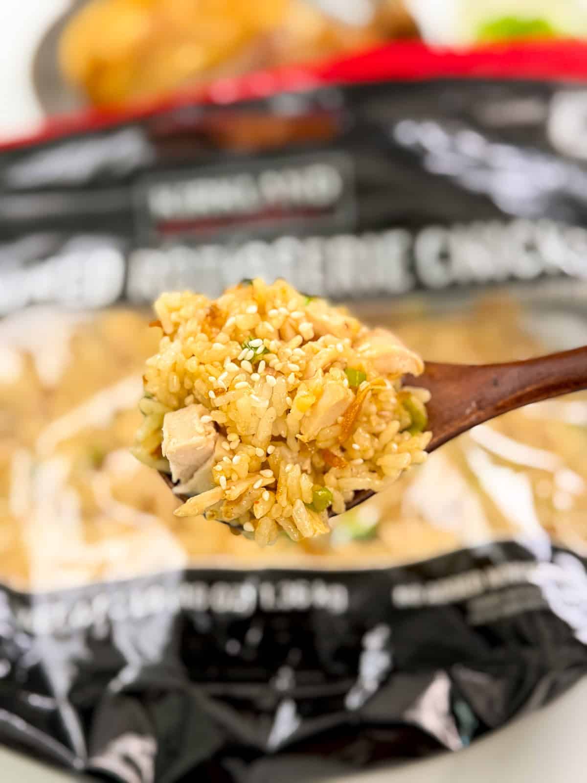 A wooden spoon holds a serving of Costco Chicken Scallion Rice with vegetables and chicken, garnished with sesame seeds. In the background, a bag labeled Kirkland Signature Rotisserie Chicken is visible.