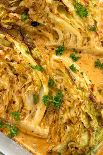 Baked cabbage wedges in a creamy, orange-colored sauce, garnished with fresh parsley, in a glass baking dish. The edges of the garlic butter melting cabbage are roasted and slightly browned.