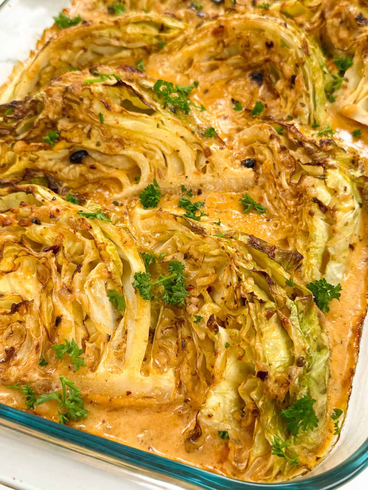 Roasted cabbage wedges covered in a creamy orange sauce and garnished with chopped fresh parsley, served in a glass baking dish.