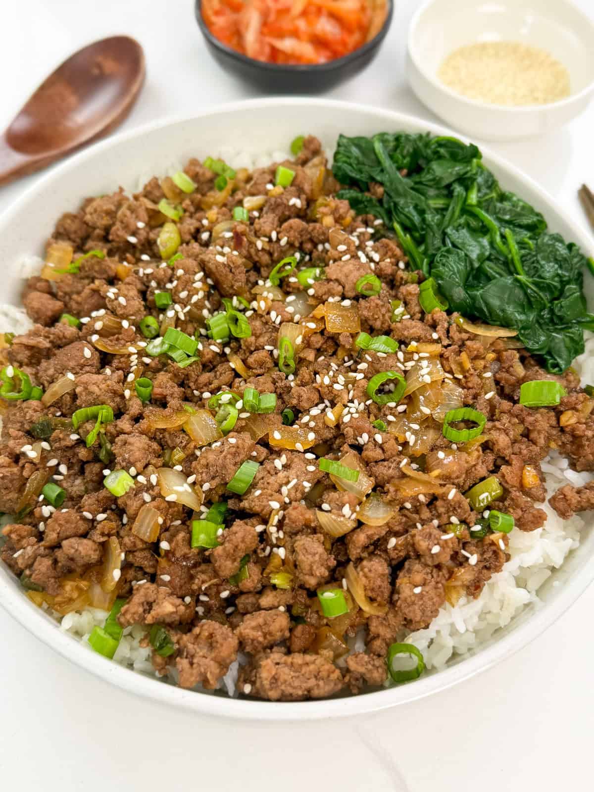 A bowl of white rice topped with Korean ground beef, chopped green onions, sautéed onions, sesame seeds, and sautéed spinach, with small dishes of kimchi and sesame seeds in the background.