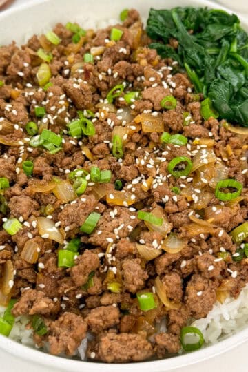 A bowl of white rice topped with seasoned Korean ground beef, sautéed onions, chopped green onions, sesame seeds, and a serving of cooked spinach on the side.
