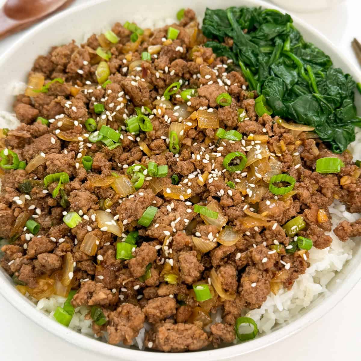 A bowl of white rice topped with seasoned Korean ground beef, sautéed onions, chopped green onions, sesame seeds, and a serving of cooked spinach on the side.