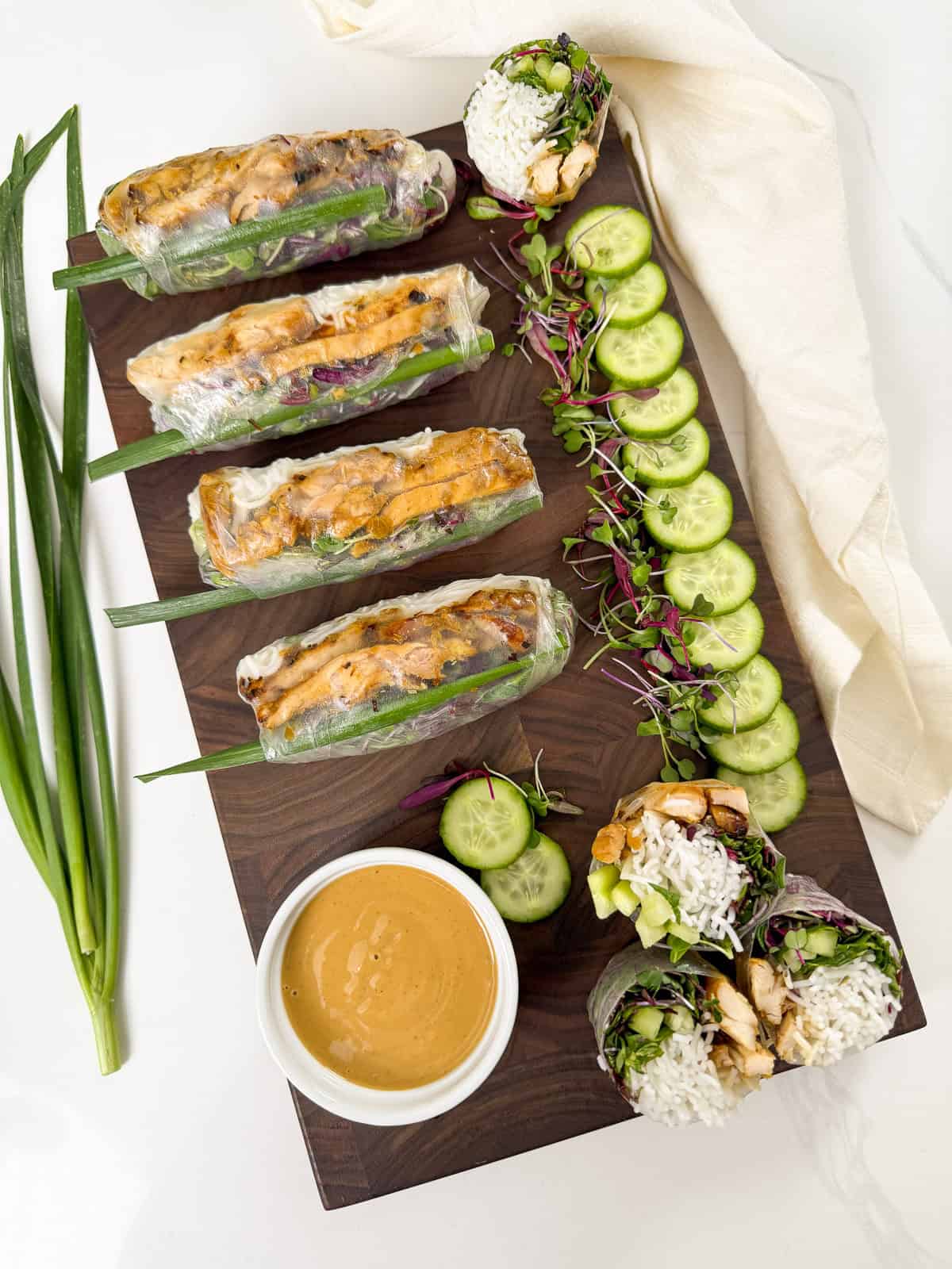 A wooden board with chicken spring rolls, sliced cucumbers, rice noodles, lettuce, green onions, and a bowl of dipping sauce. Some rolls are cut open, showing the filling. A white napkin sits to the side.
