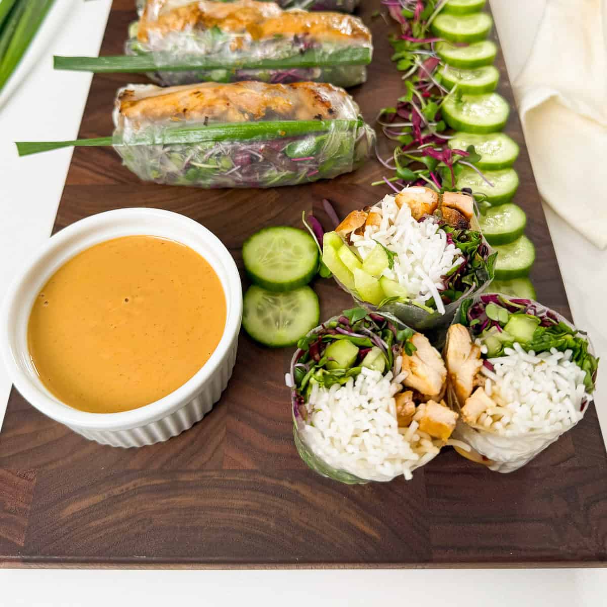 A wooden board with fresh spring rolls filled with rice, chicken, cucumber, and greens, sliced cucumbers, and a small white ramekin of peanut dipping sauce.