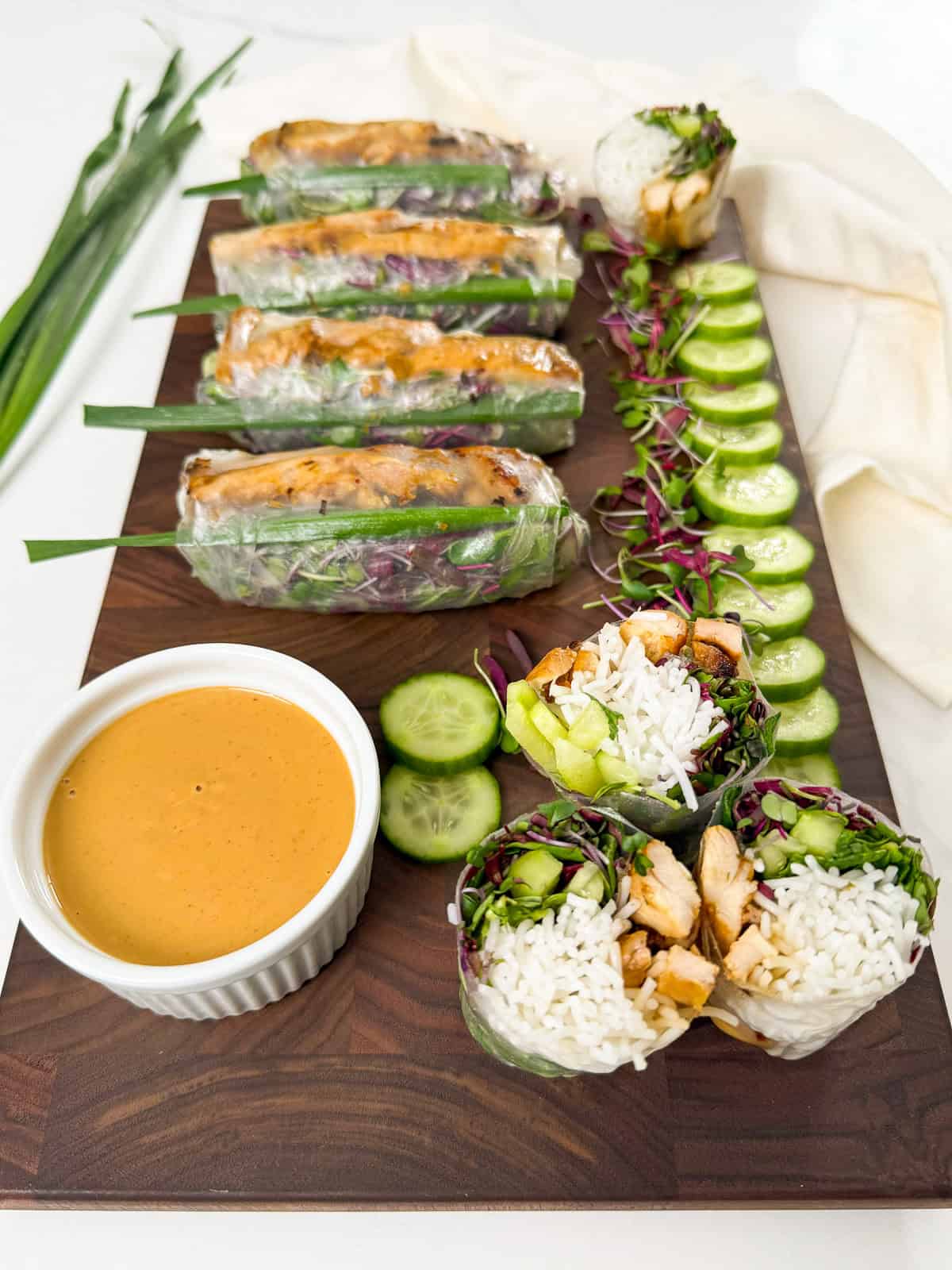 A wooden board with neatly arranged chicken and vegetable rice paper rolls, sliced cucumber, a ramekin of dipping sauce, and garnished with green onions. One roll is cut open to show the fresh ingredients inside.