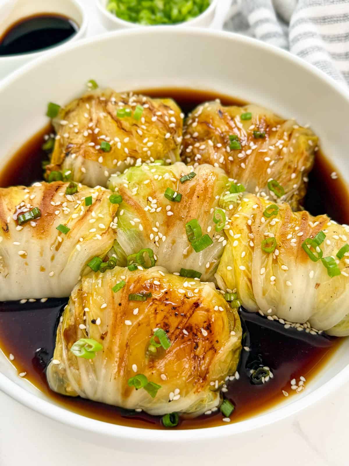 Napa cabbage rolls in a bowl with sweet soy sauce with sesame seeds, green onions, and chili oil main view