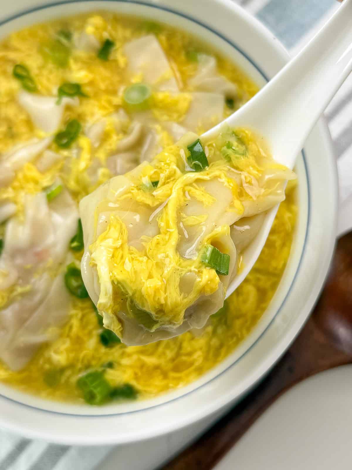 A close-up of a spoon holding a dumpling above a bowl of egg drop soup with green onions, showing the soup’s yellow egg ribbons and bits of dumpling filling.