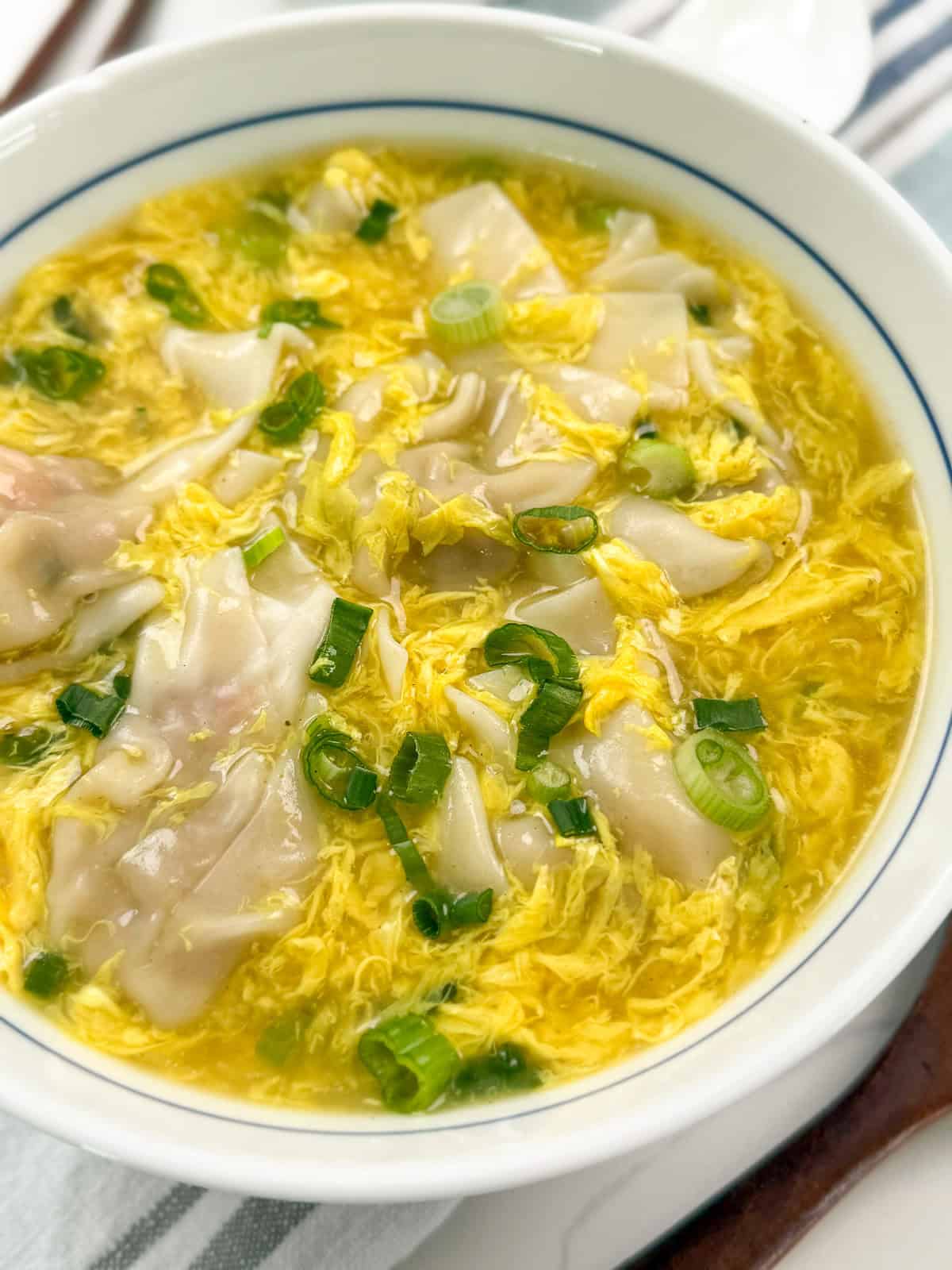 A bowl of wonton egg drop soup, featuring dumplings, wisps of cooked egg, and chopped green onions in a clear yellow broth.