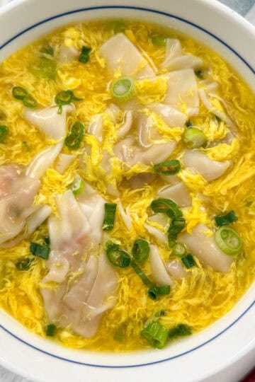 A bowl of wonton soup with yellow egg ribbons, garnished with chopped green onions, served in a white bowl with blue trim.