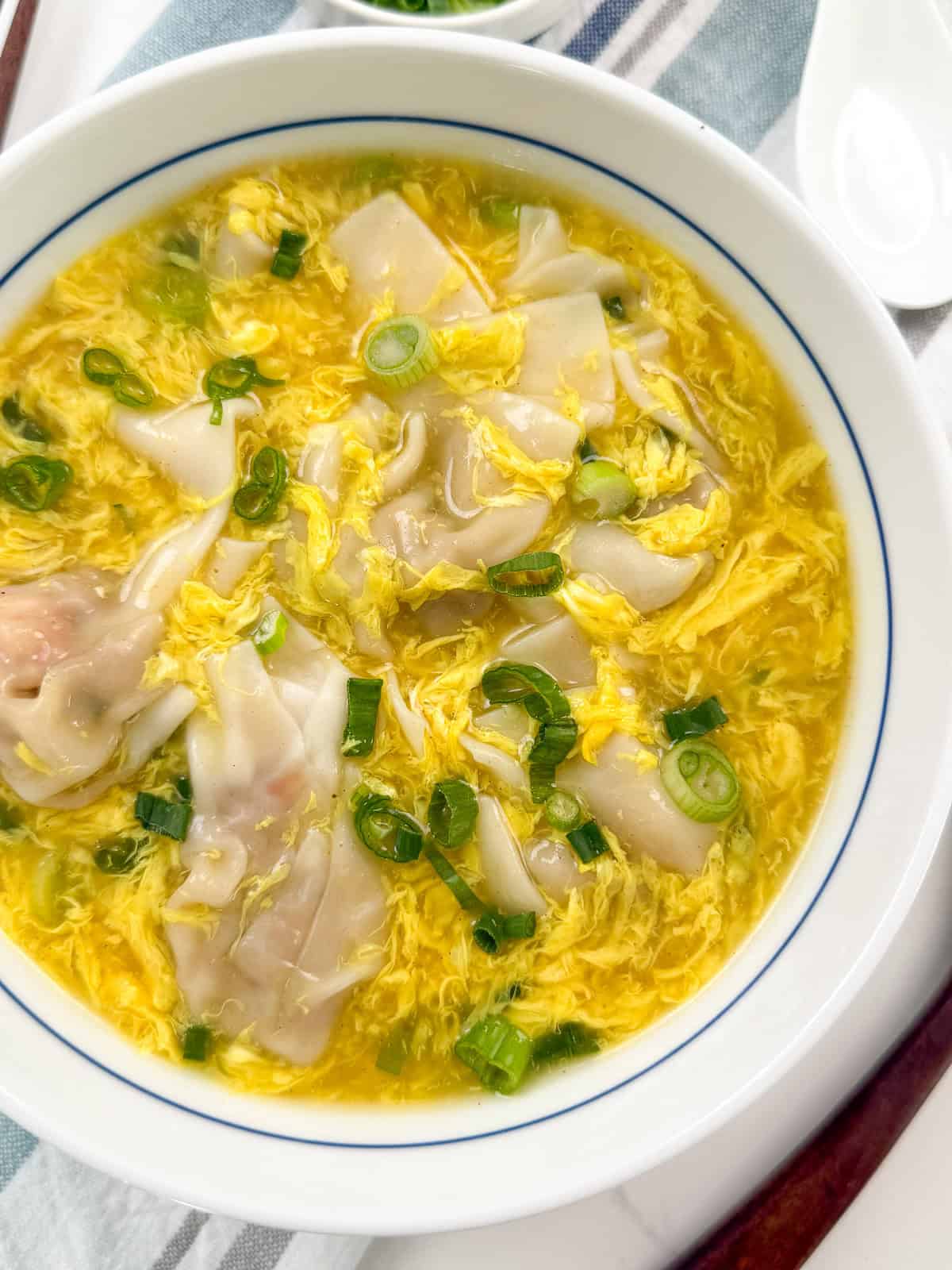 A bowl of egg drop soup with dumplings, garnished with chopped green onions, sits on a striped cloth with a white spoon nearby. The soup has a yellow broth with visible cooked egg ribbons.