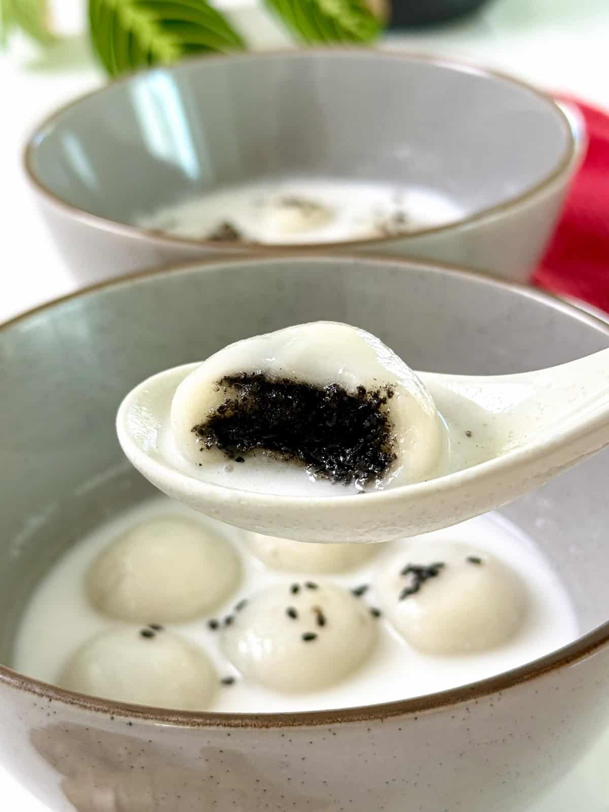 A close-up of a spoon holding a Black Sesame Glutinous Rice Ball (Tang Yuan) with rich black sesame filling, above a bowl of sweet soup containing more Tang Yuan. Another bowl is visible in the background.
