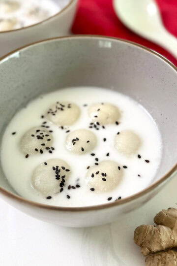 A bowl of Black Sesame Glutinous Rice Balls (Tang Yuan) in coconut milk, topped with black sesame seeds. A ceramic spoon, a red cloth, and a piece of fresh ginger are nearby on a white surface.