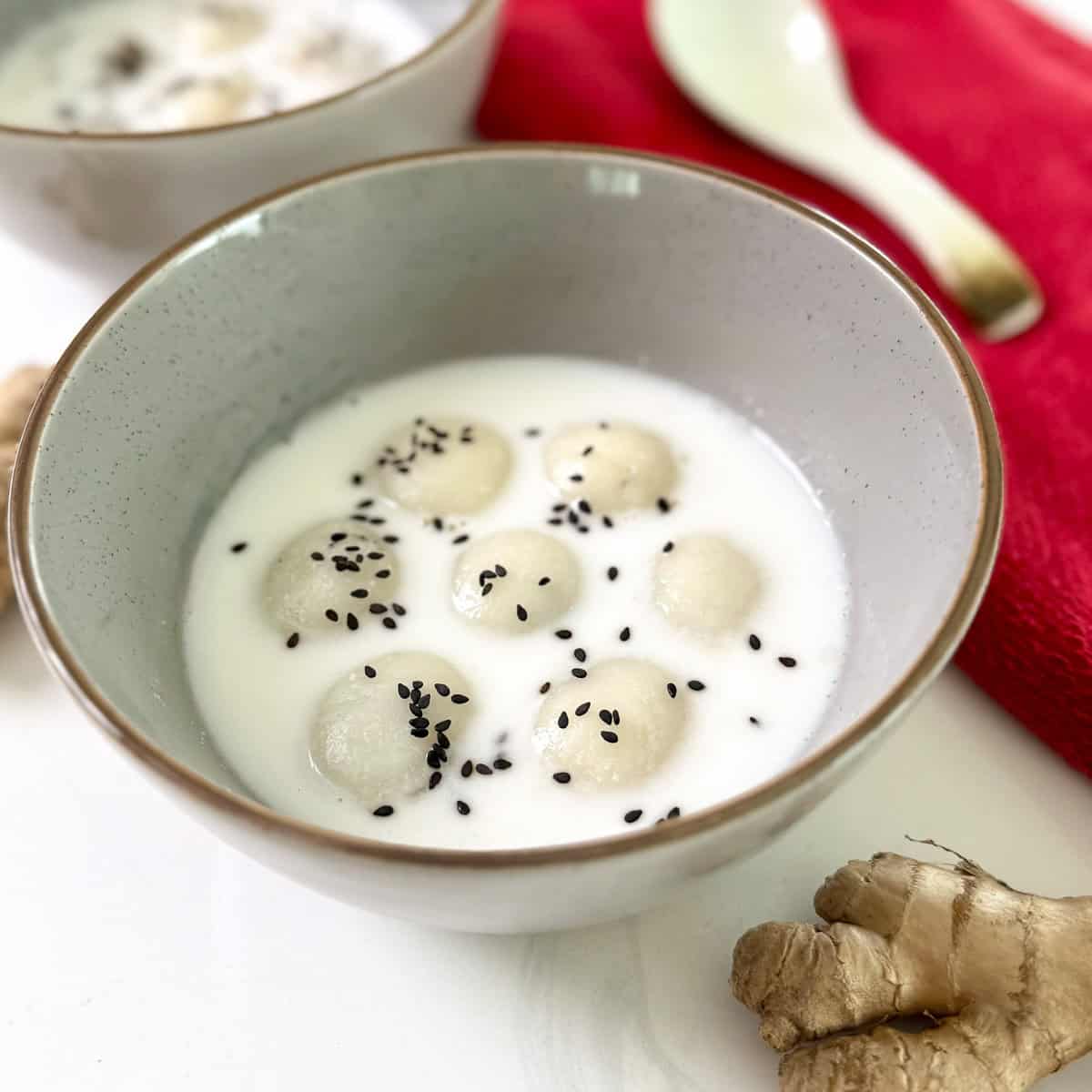 A bowl of Black Sesame Glutinous Rice Balls (Tang Yuan) in coconut milk, topped with black sesame seeds. A ceramic spoon, a red cloth, and a piece of fresh ginger are nearby on a white surface.