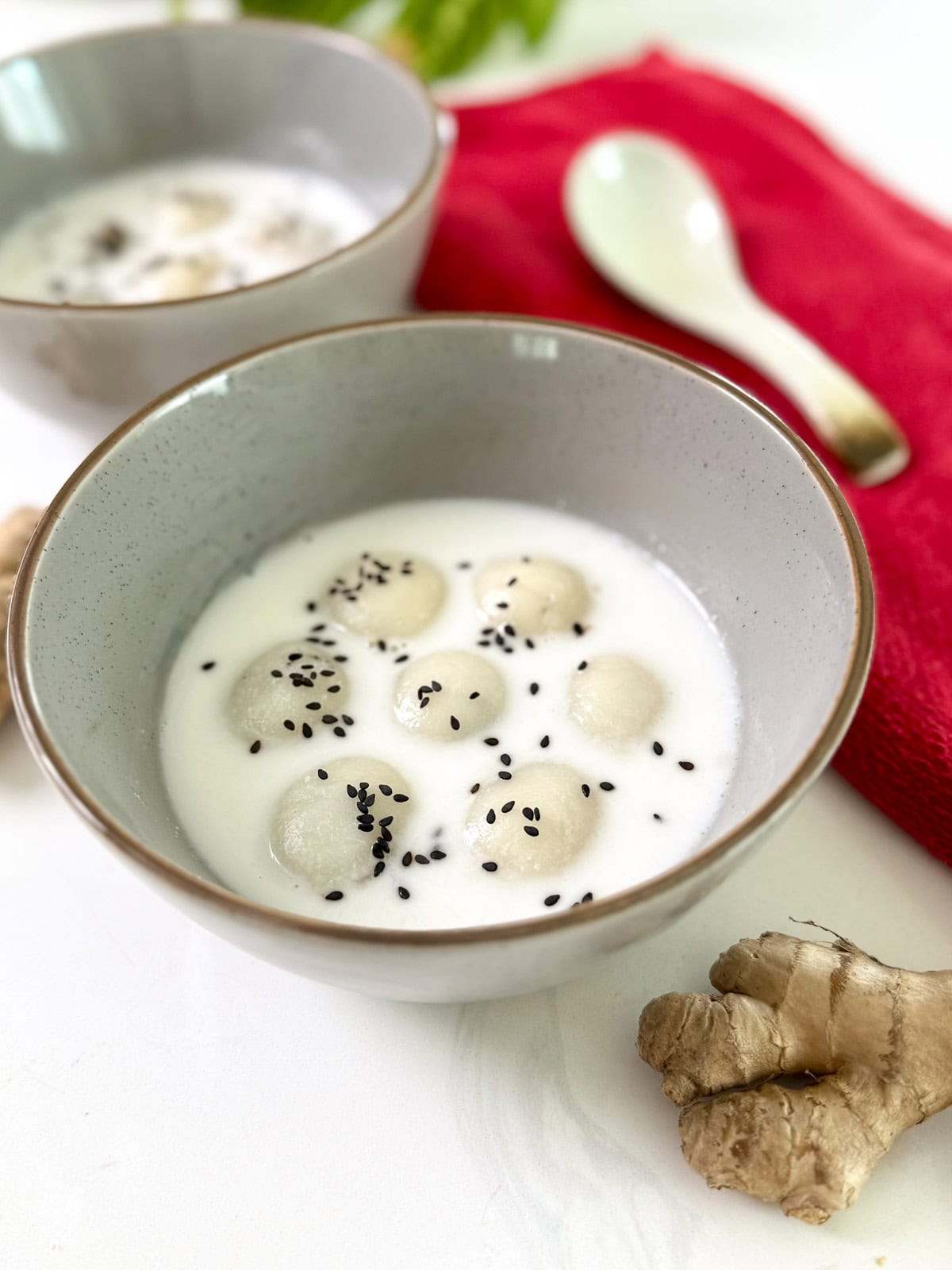 A bowl of Black Sesame Glutinous Rice Balls (Tang Yuan) in coconut milk, topped with black sesame seeds. Another bowl, a spoon, a red cloth, and fresh ginger are nearby on a white surface.
