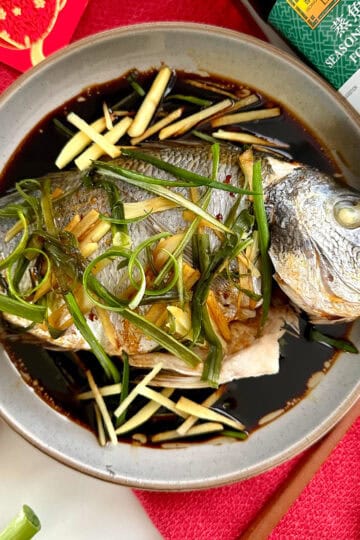 A whole Cantonese Steamed Fish, garnished with julienned ginger and green onions, sits in a shallow bowl of soy sauce. Chopsticks, fresh ginger, a red cloth, and a soy sauce bottle are placed nearby.