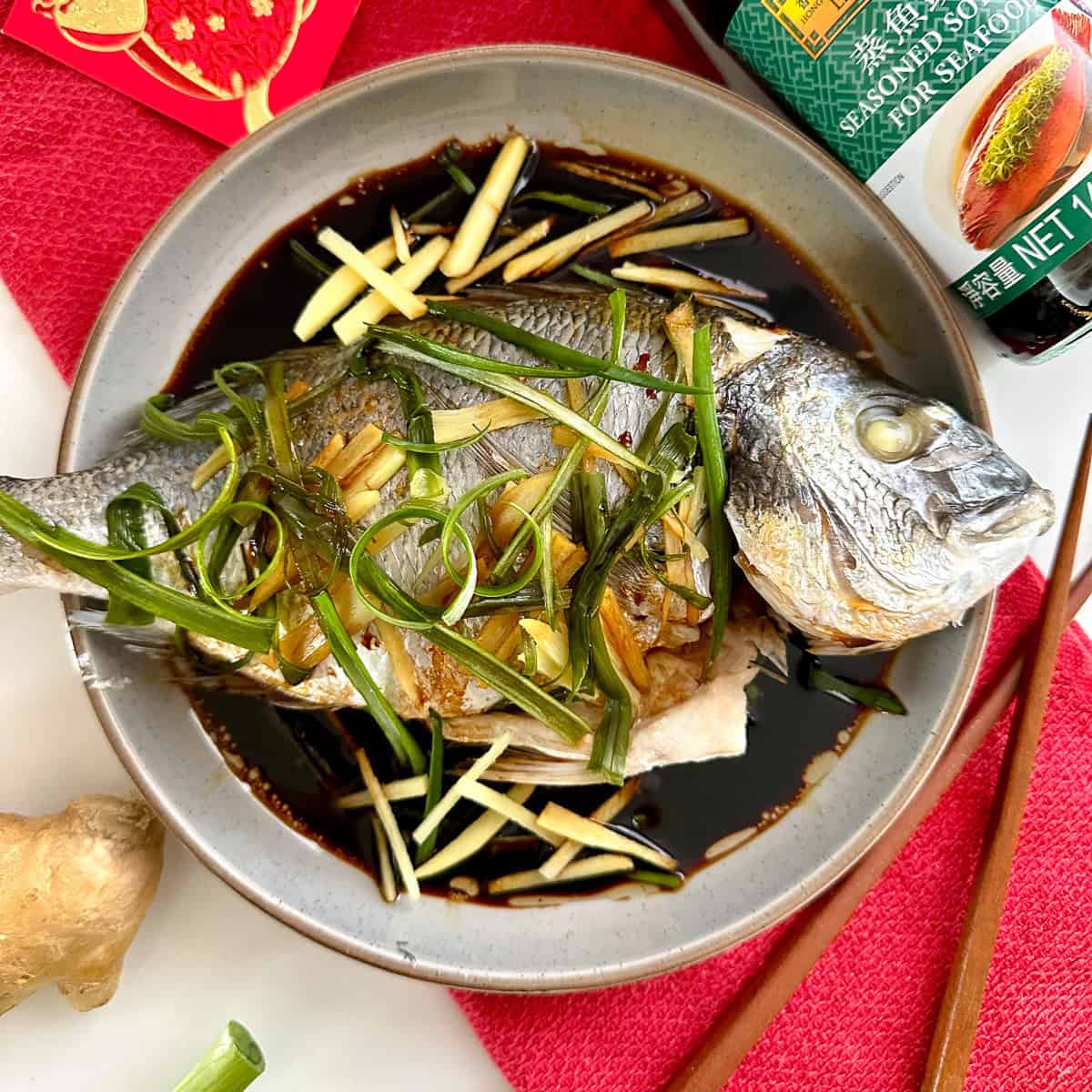 A whole Cantonese Steamed Fish, garnished with julienned ginger and green onions, sits in a shallow bowl of soy sauce. Chopsticks, fresh ginger, a red cloth, and a soy sauce bottle are placed nearby.