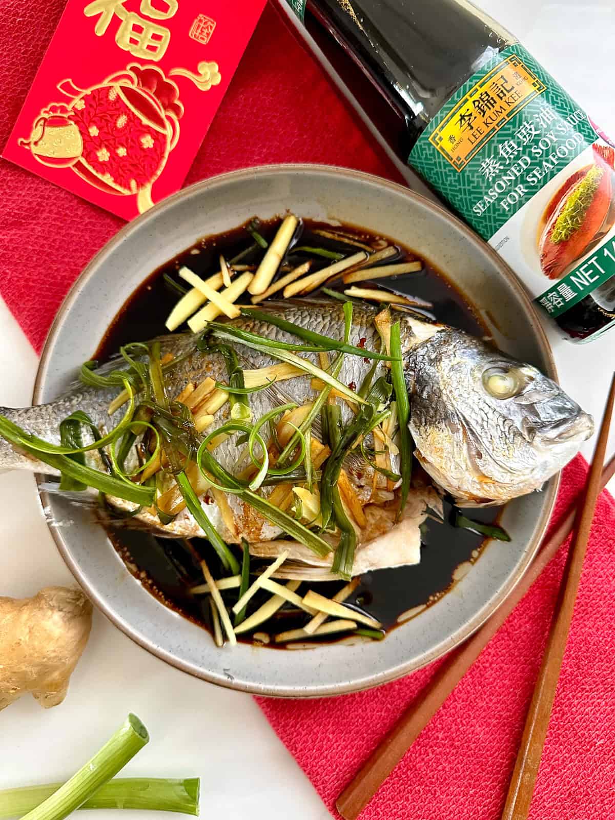 A Cantonese steamed fish topped with julienned ginger and scallions sits in a soy sauce-based broth in a gray bowl. Nearby are chopsticks, a soy sauce bottle, a red envelope, and a piece of fresh ginger.
