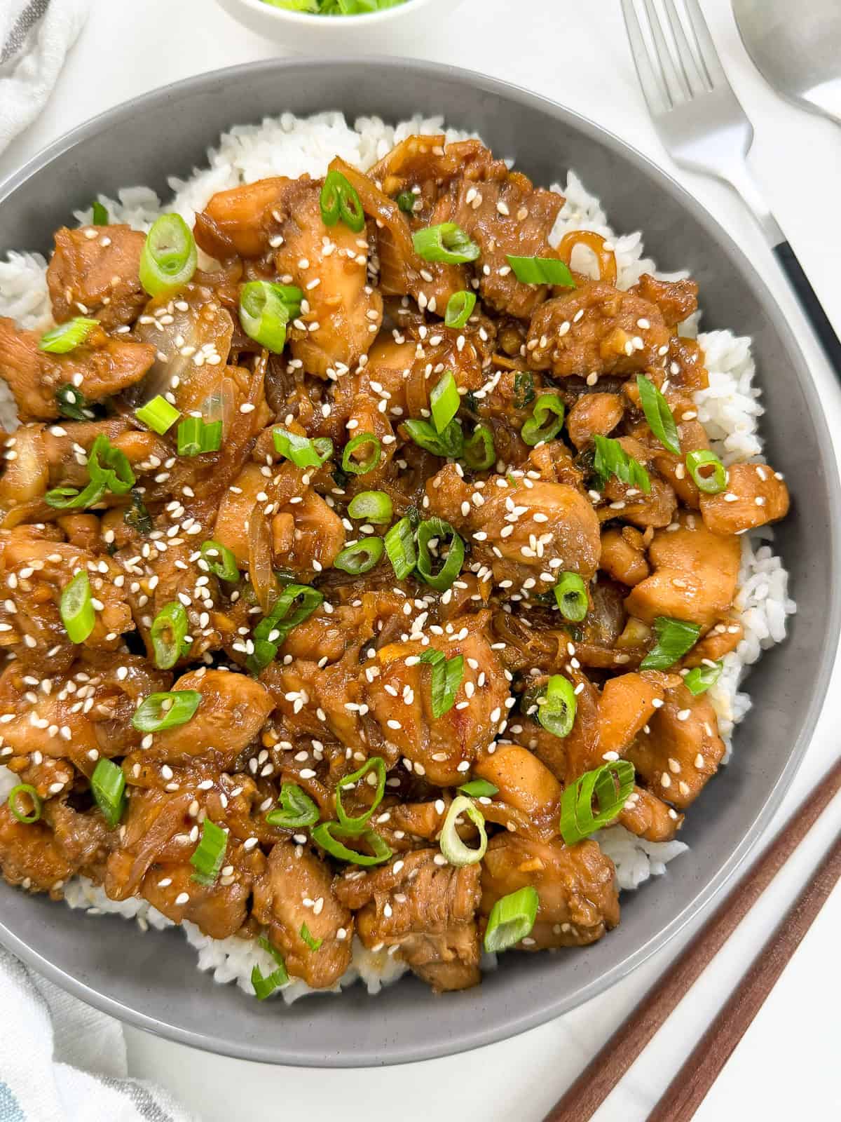 A bowl of white rice topped with Chicken Bulgogi (Korean BBQ Chicken), glazed to perfection and garnished with sliced green onions and sesame seeds. A spoon and fork are visible beside the bowl.