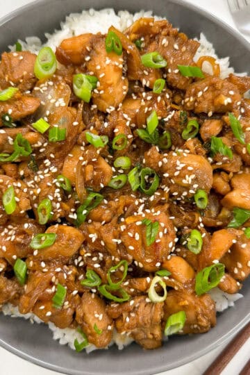 A bowl of white rice topped with glazed Chicken Bulgogi (Korean BBQ Chicken) pieces, garnished with sliced green onions and sprinkled with sesame seeds. A fork and knife are visible beside the bowl.