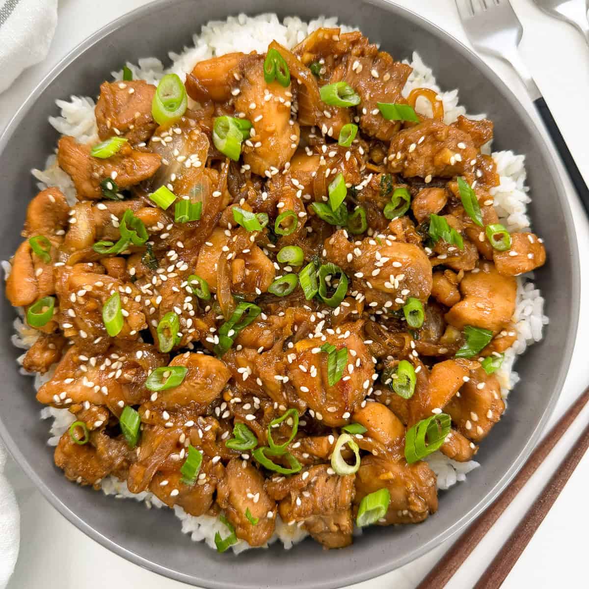A bowl of white rice topped with glazed Chicken Bulgogi (Korean BBQ Chicken) pieces, garnished with sliced green onions and sprinkled with sesame seeds. A fork and knife are visible beside the bowl.