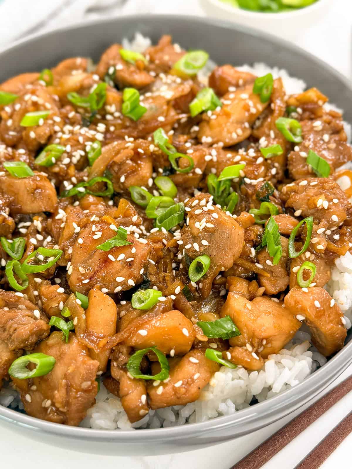 A bowl of white rice topped with glazed Chicken Bulgogi (Korean BBQ Chicken) pieces, sliced green onions, and sprinkled sesame seeds.