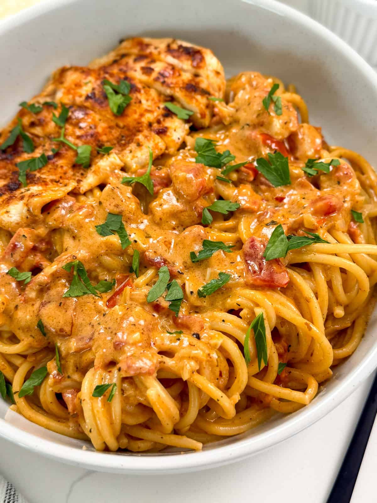 A bowl of Creamy Cajun Chicken Pasta topped with a rich tomato sauce, garnished with chopped parsley, and served with a piece of seasoned, grilled chicken breast.