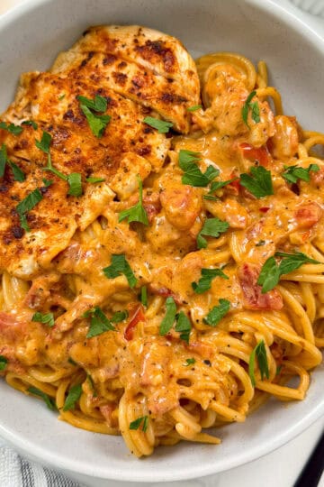 A bowl of Creamy Cajun Chicken Pasta features spaghetti topped with a rich tomato sauce, garnished with chopped parsley, and served with a grilled chicken breast.
