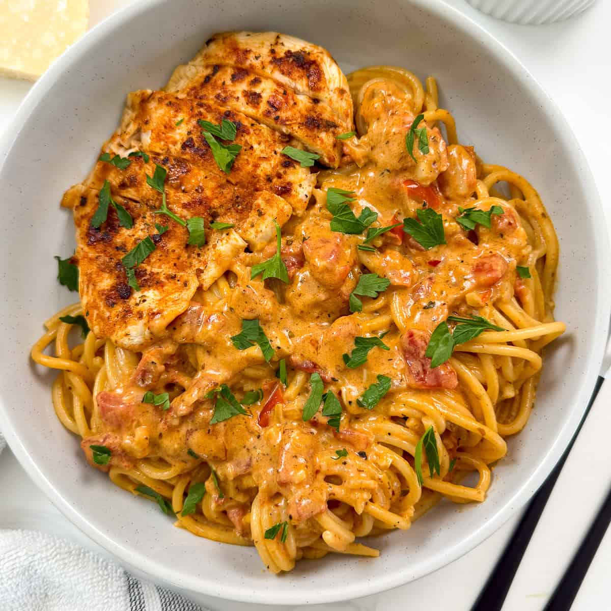 A bowl of Creamy Cajun Chicken Pasta features spaghetti topped with a rich tomato sauce, garnished with chopped parsley, and served with a grilled chicken breast.