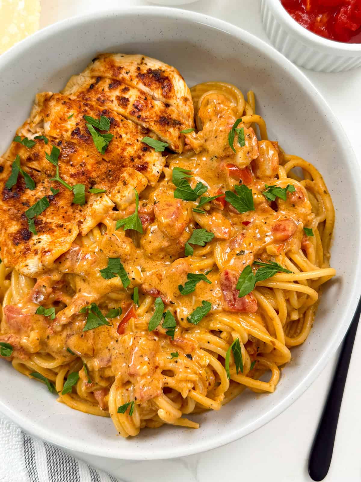 A bowl of spaghetti topped with a creamy tomato sauce and garnished with chopped parsley, served with a grilled chicken breast on the side—a delicious twist on Creamy Cajun Chicken Pasta.