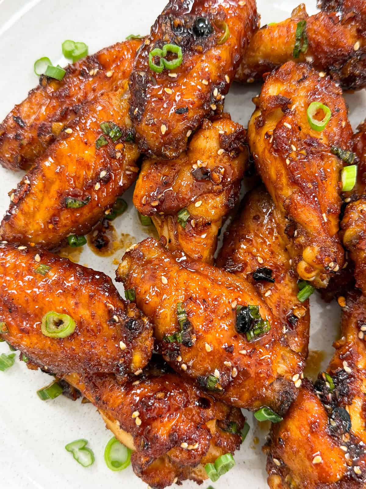 A plate of crispy honey chili oil chicken wings garnished with chopped green onions and sesame seeds. The wings have a shiny, caramelized coating and are arranged closely together on a white plate.