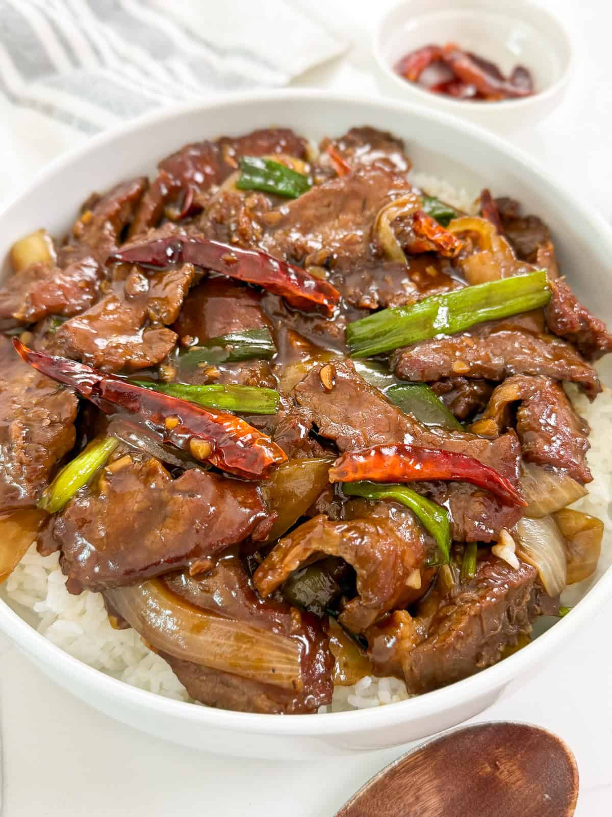 A bowl of white rice topped with Mongolian Beef Recipe: tender stir-fried beef, green onions, onions, and dried red chilies in a glossy sauce, with a small bowl of extra chilies in the background.