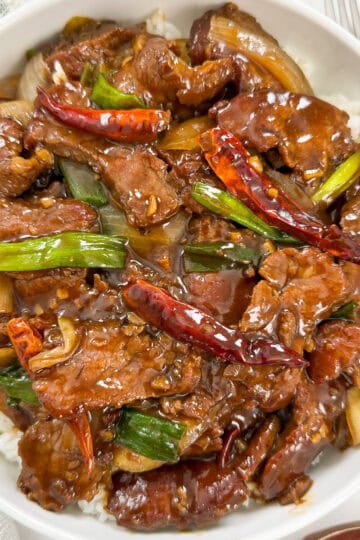 A bowl of Mongolian Beef Recipe stir-fried with green onions, dried red chilies, and onions in a glossy brown sauce, served over white rice.