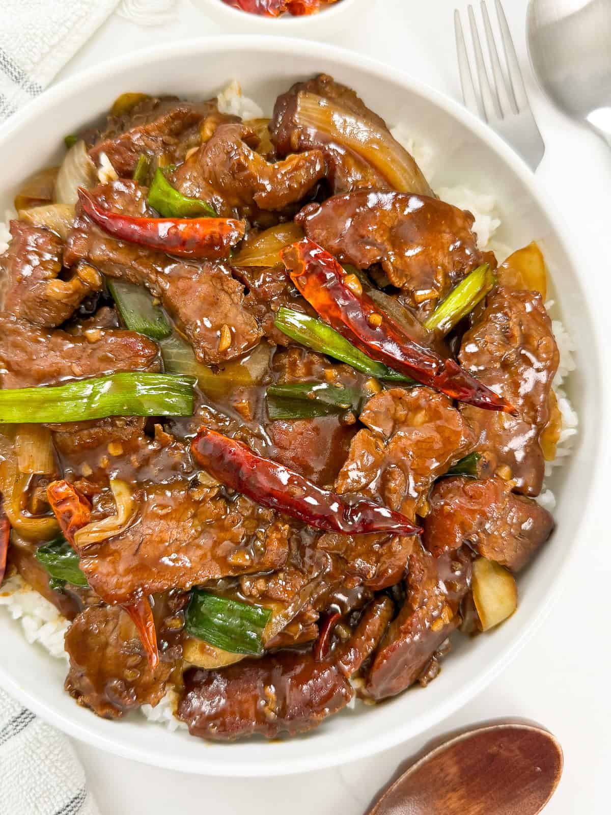 A bowl of spicy Mongolian Beef stir-fry with red chili peppers, green onions, and onions served over steamed white rice on a white table, with a spoon and fork nearby—a classic Mongolian Beef Recipe favorite.