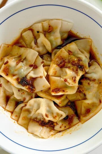 A white bowl filled with Pork and Chive Dumplings, topped with chili oil and spices, shows a glistening, flavorful sauce over perfectly folded dumpling wrappers.