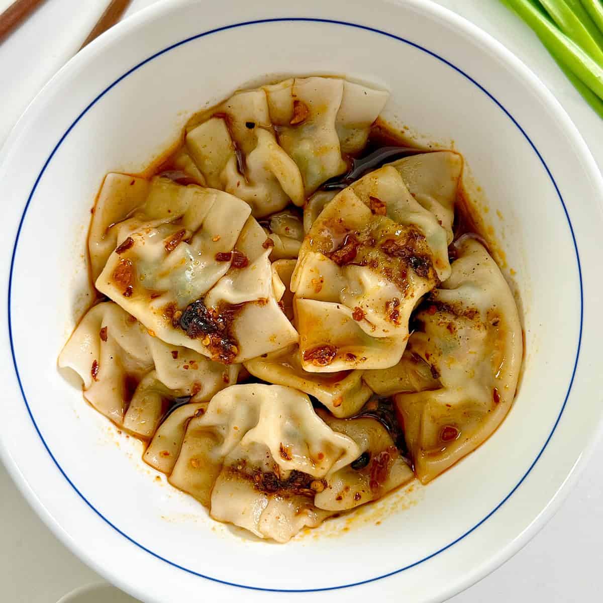 A white bowl filled with Pork and Chive Dumplings, topped with chili oil and spices, shows a glistening, flavorful sauce over perfectly folded dumpling wrappers.
