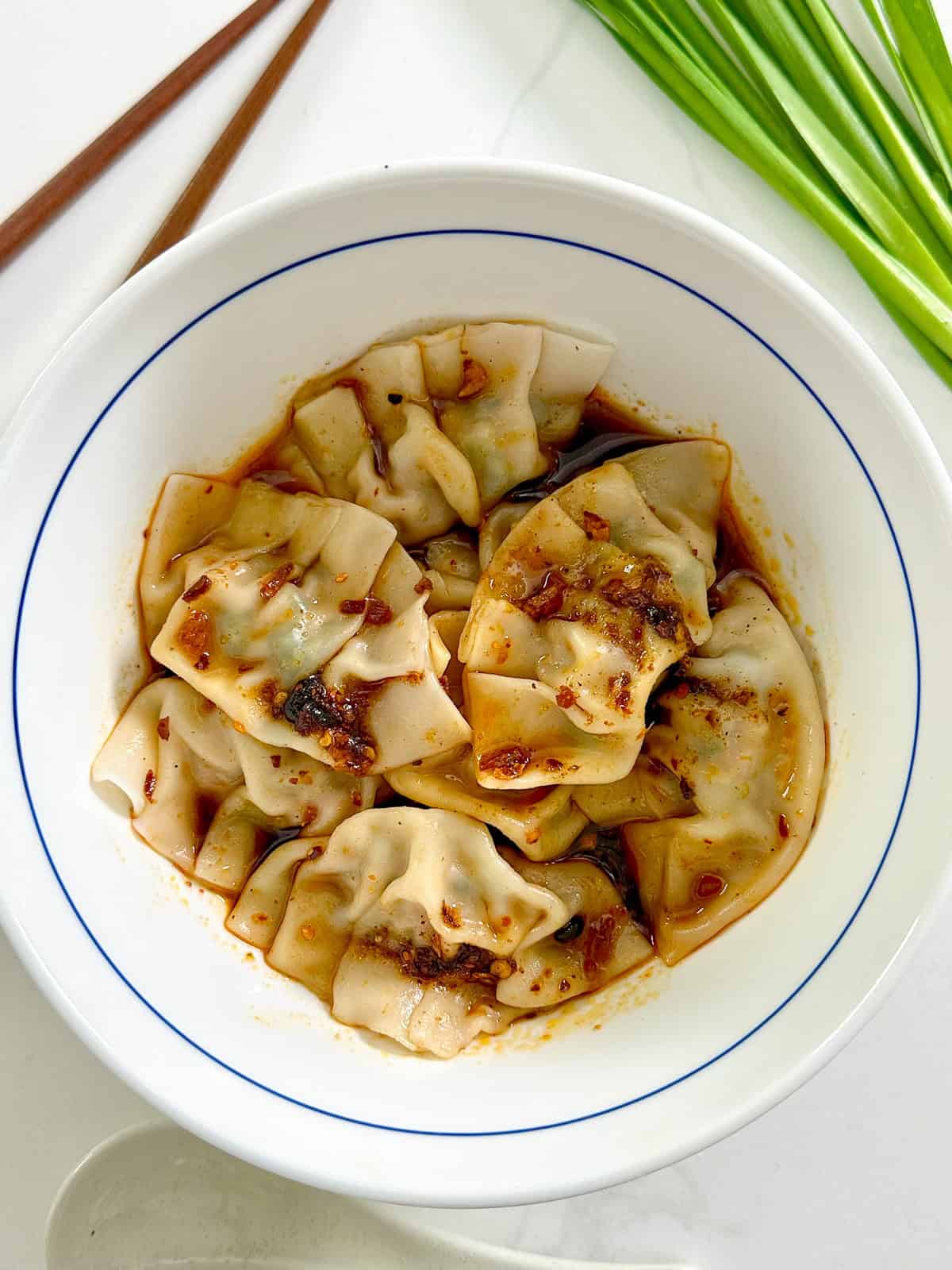 A white bowl filled with Pork and Chive Dumplings topped with chili oil sauce, placed on a white surface next to chopsticks and fresh green onions.