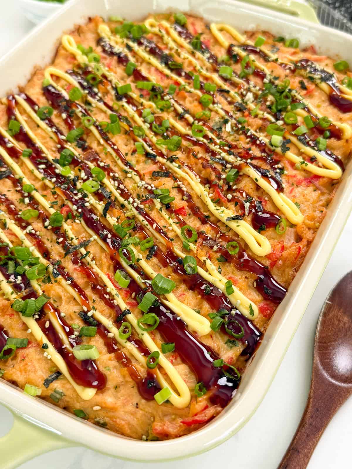 A close-up of a Salmon Sushi Bake casserole topped with zigzag drizzles of mayonnaise and dark sauce, garnished with chopped green onions, sesame seeds, and herbs, in a rectangular dish with a wooden spoon beside it.