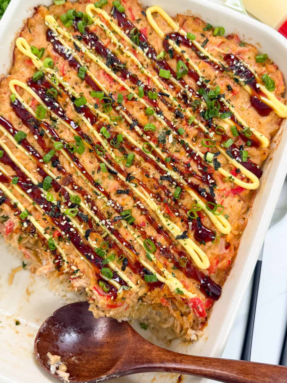 A Salmon Sushi Bake casserole topped with drizzles of mayonnaise and dark sauce, garnished with chopped green onions and sesame seeds. A wooden spoon rests in the dish, showing a serving has been scooped out.
