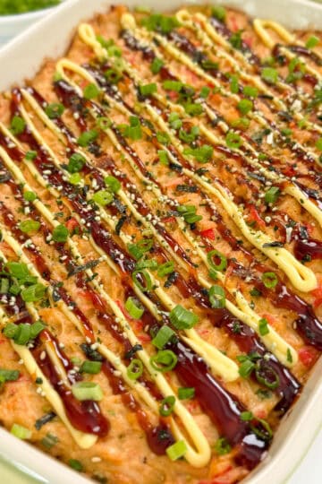 A Salmon Sushi Bake casserole topped with drizzles of brown and yellow sauces, sprinkled with chopped green onions and sesame seeds, in a white rectangular dish.