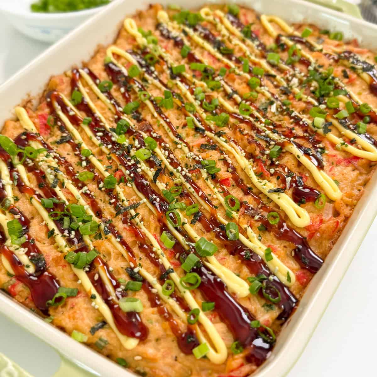 A Salmon Sushi Bake casserole topped with drizzles of brown and yellow sauces, sprinkled with chopped green onions and sesame seeds, in a white rectangular dish.