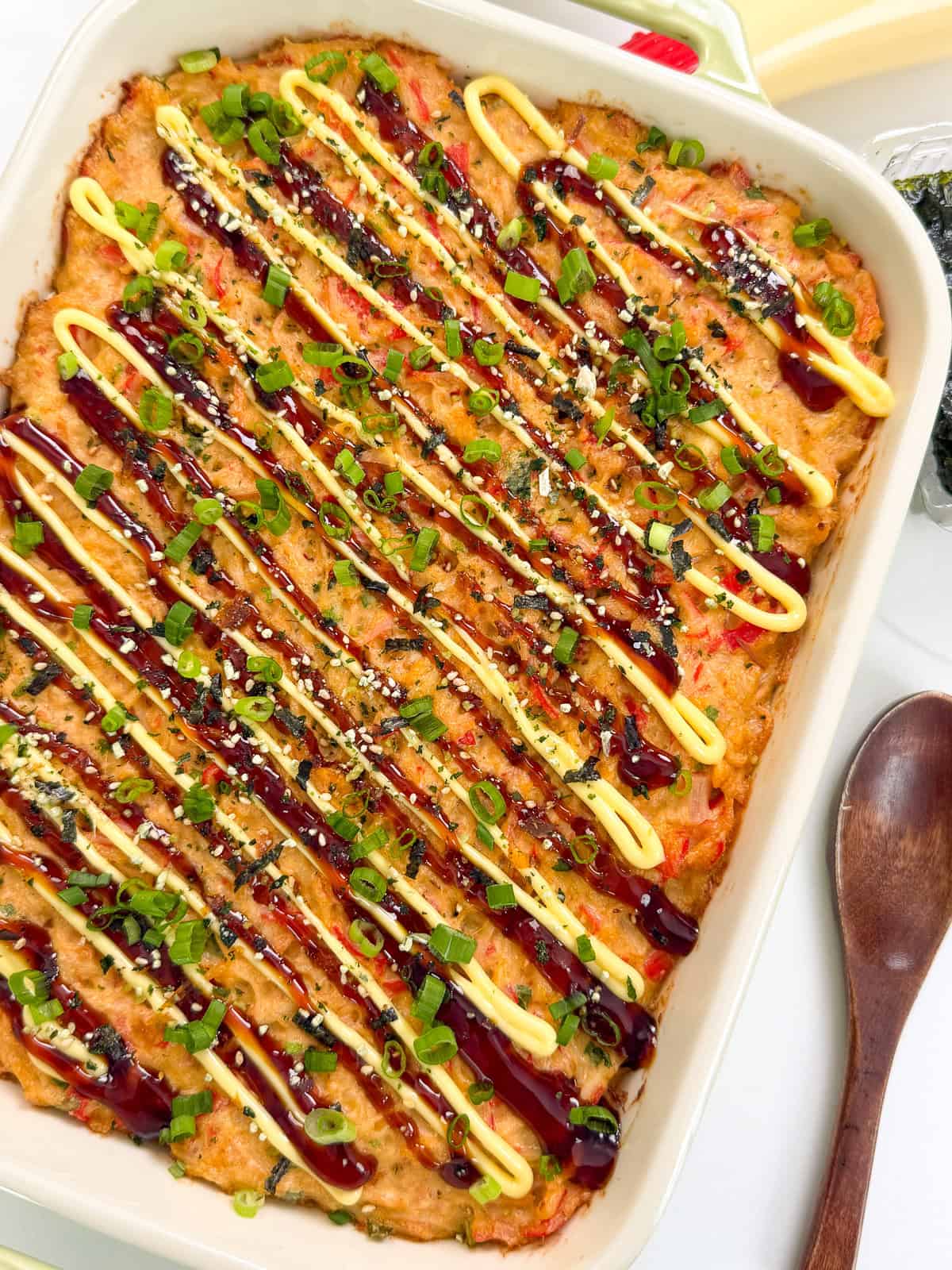 A Salmon Sushi Bake casserole dish filled with baked sushi rice, topped with zigzag drizzles of creamy mayo and dark sauce, sprinkled with chopped green onions and sesame seeds, with a wooden spoon and nori sheets beside it.