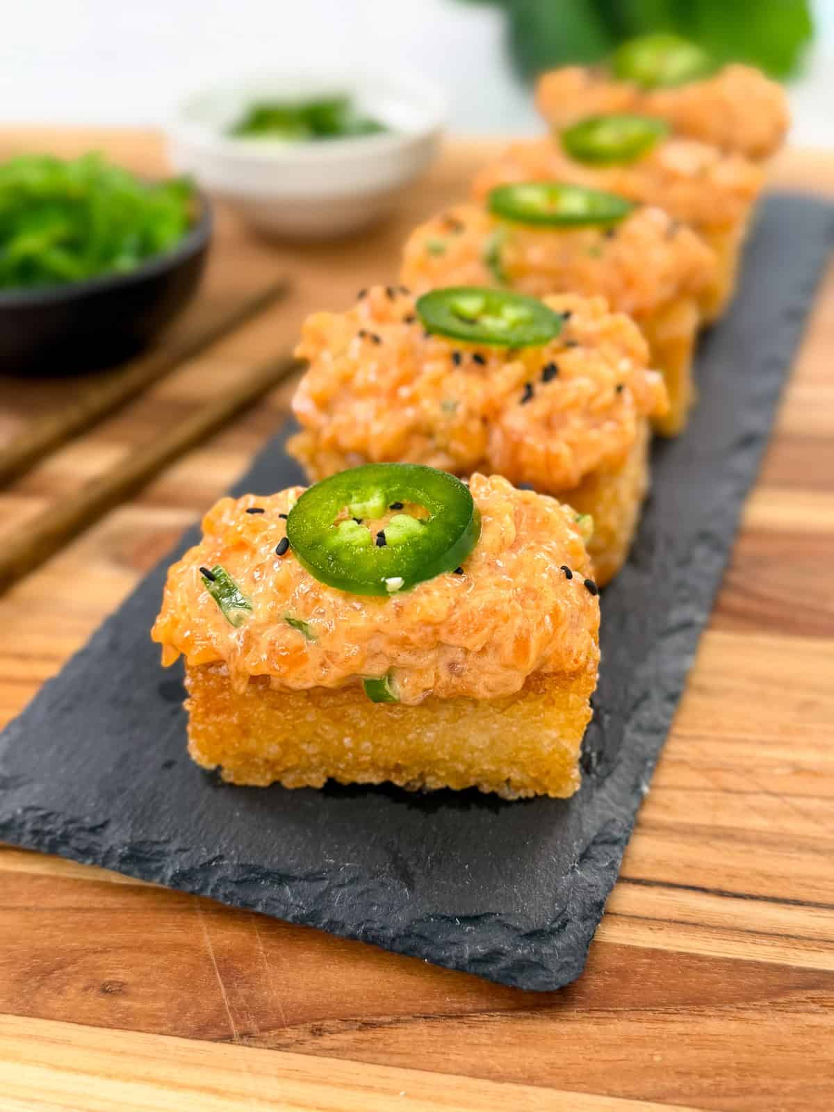 Four pieces of Spicy Salmon Crispy Rice topped with a spicy salmon mixture and sliced jalapeño are arranged in a row on a slate serving tray, with small bowls of garnish in the background.